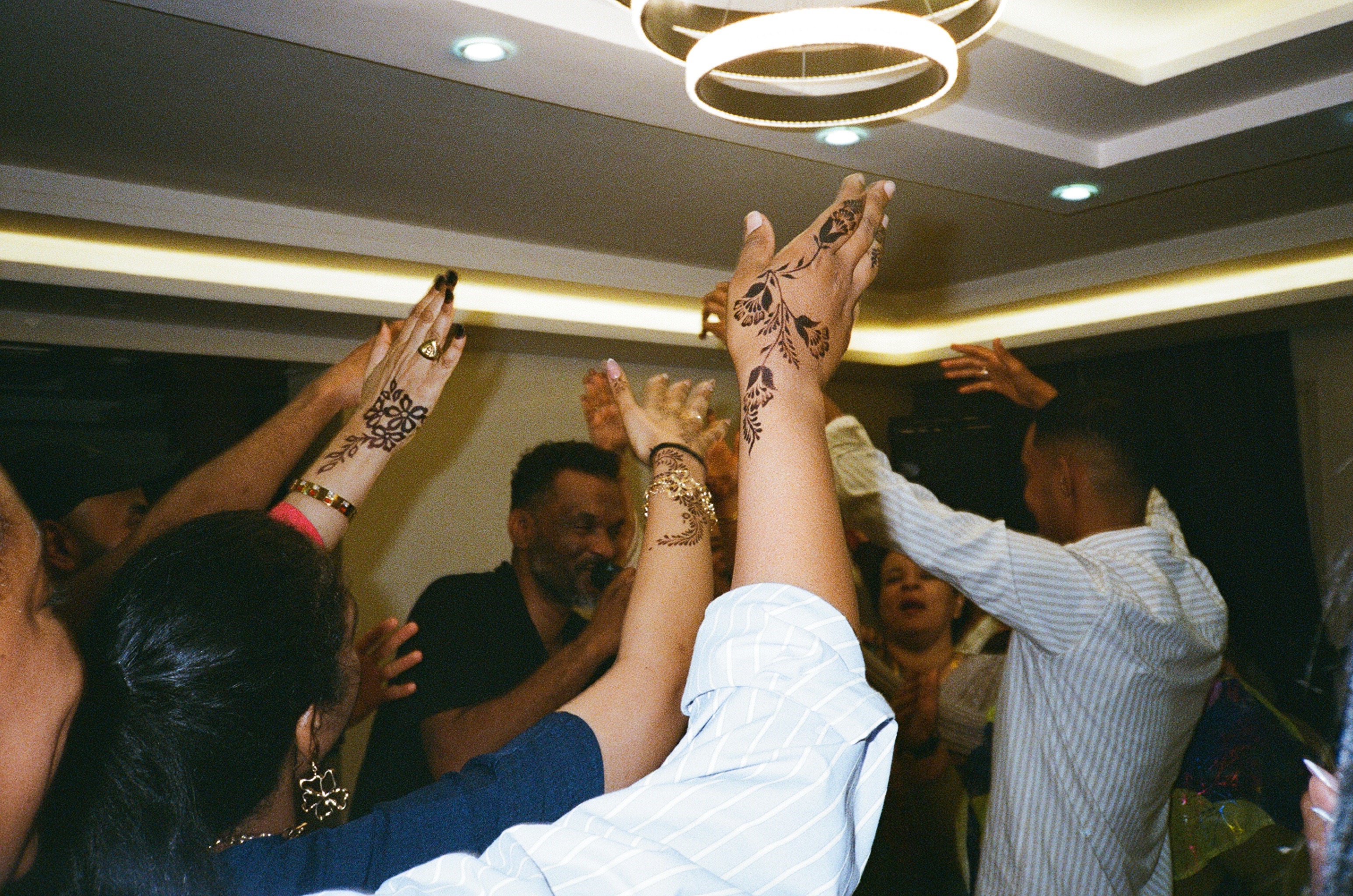 A photo of people dancing together. Several arms are raised in the air. Some hands are decorated with henna.